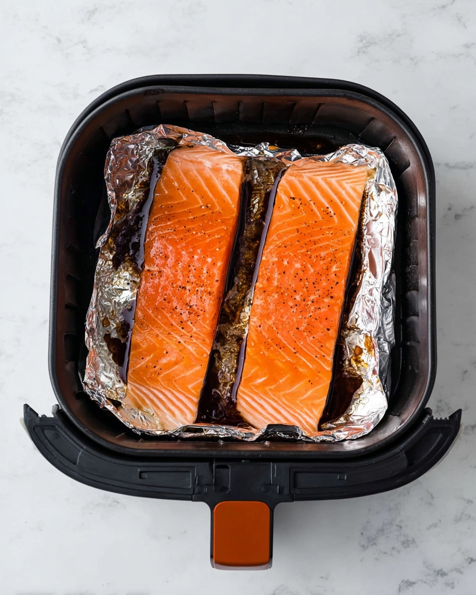 Two long, raw salmon fillets with a bright orange color and faint white lines lay flat side by side inside a black air fryer basket. The basket is lined with crinkled silver foil that covers the sides, catching some dark soy sauce or marinade pooling underneath the fish. The salmon’s surface looks smooth and moist with small black pepper specks scattered across. The air fryer basket has a black body with an orange handle, resting on a white marbled surface photo taken with an iphone --ar 4:5 --v 7