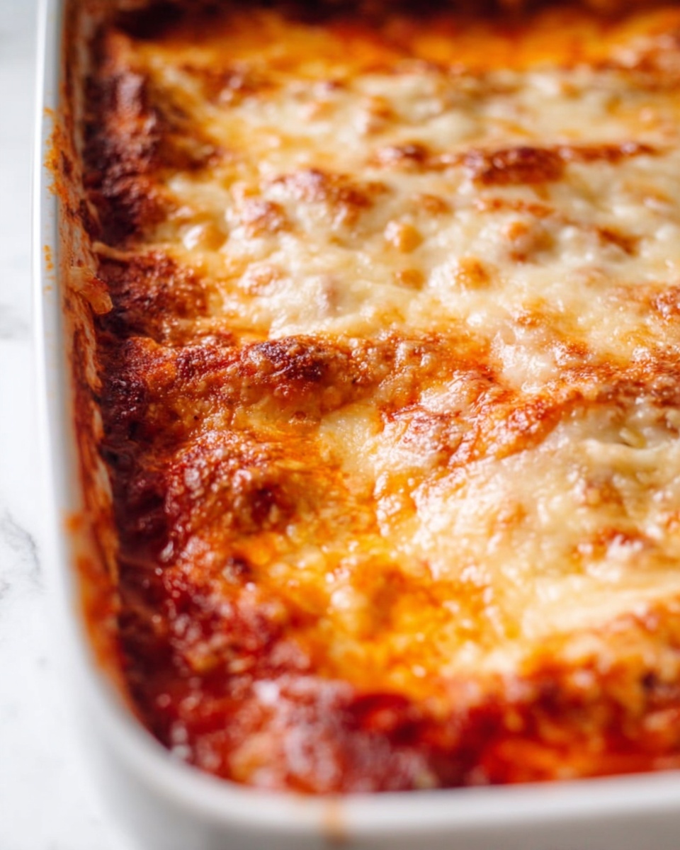 The image shows a close-up of a baked dish in a white baking tray with reddish-orange sauce and a thick layer of melted cheese on top. The melted cheese is golden brown in some spots, with a bubbly and slightly crispy texture. The sauce underneath is visible around the edges, showing a chunky and moist consistency with a rich tomato color. The white marbled surface beneath the tray is softly blurred, keeping focus on the cheesy baked dish photo taken with an iphone --ar 4:5 --v 7