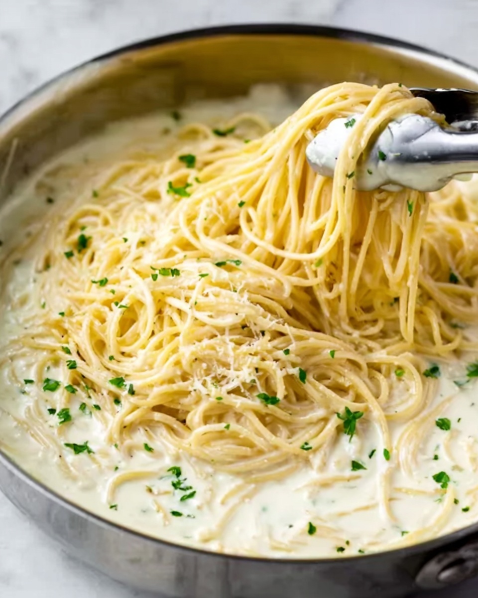 This image shows a close-up of a pan filled with creamy Alfredo pasta. The bottom layer is the white creamy sauce, thick and smooth, covering the base of the pan. On top of this sauce is a layer of cooked spaghetti noodles, light yellow in color, with some noodles lifted slightly by metal tongs held by a woman's hand. Small green parsley pieces are sprinkled evenly across the noodles adding a touch of color. The background is a white marbled texture. Photo taken with an iphone --ar 4:5 --v 7