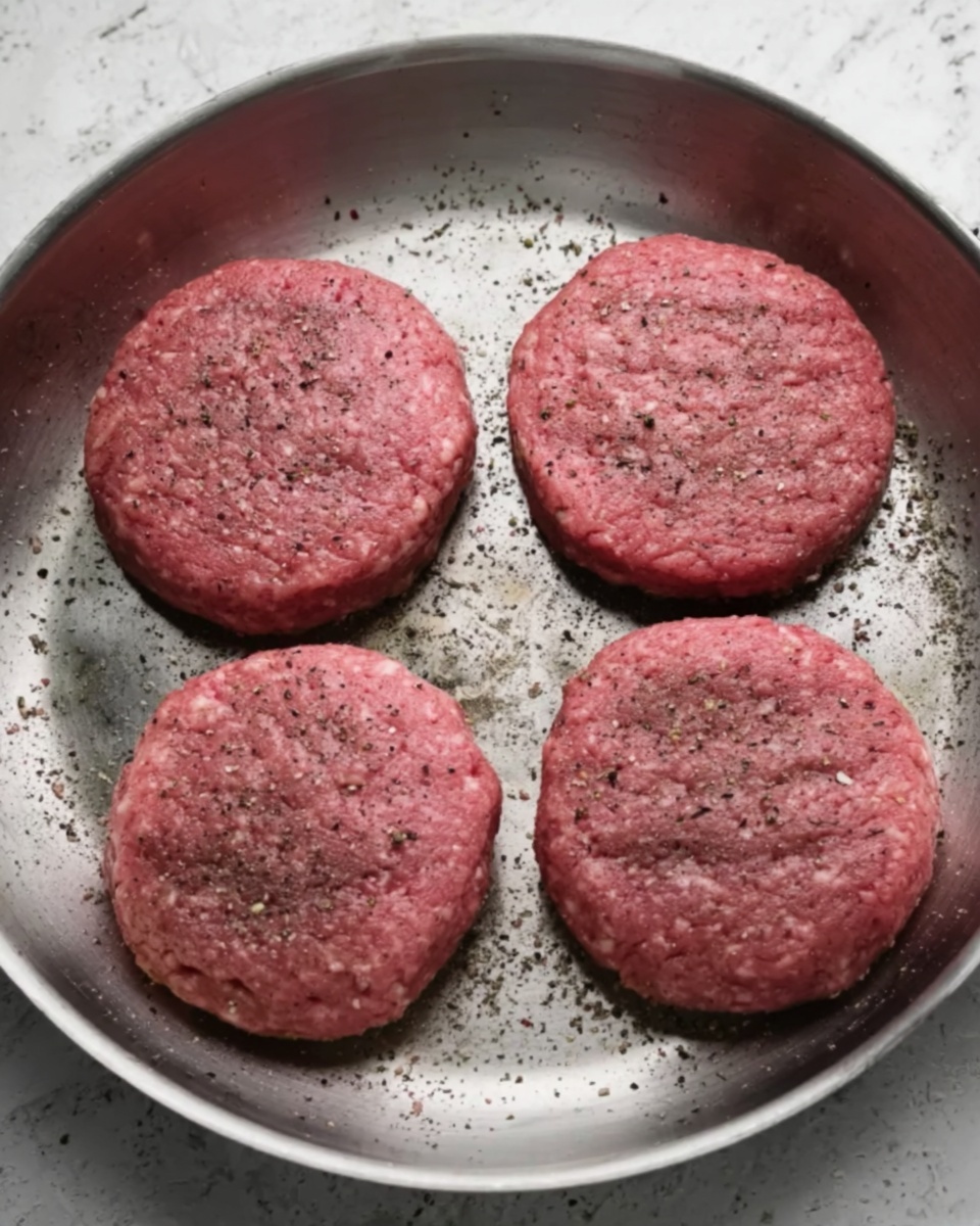 A round silver pan with four evenly spaced raw hamburger patties inside. Each patty is pink with a slightly rough texture and sprinkled with black pepper. The pan shows some small scorch marks and has a shiny metal surface. The background is a white marbled texture photo taken with an iphone --ar 4:5 --v 7