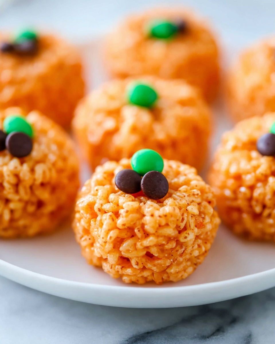 The image shows several small pumpkin-shaped treats made from orange-colored crispy rice balls. Each ball is round with a bumpy texture from the rice cereal and has a single small green candy on top representing the pumpkin stem, alongside one dark brown chocolate chip near the center. The treats are placed closely together on a white plate, which is set on a white marbled surface. The focus is on the front pumpkin treat, making the others softly blurred in the background, creating a cozy and inviting look. photo taken with an iphone --ar 4:5 --v 7