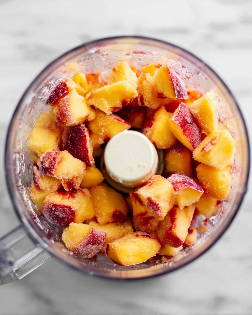A clear blender bowl filled with chunks of yellow-orange peaches with red-purple skin pieces scattered throughout, the peach pieces showing soft and slightly wet texture with some frost or ice crystals visible on the edges. The blender is placed on a white marbled surface, viewed from above, and the central white blender blade holder is visible in the middle of the fruit. photo taken with an iphone --ar 4:5 --v 7