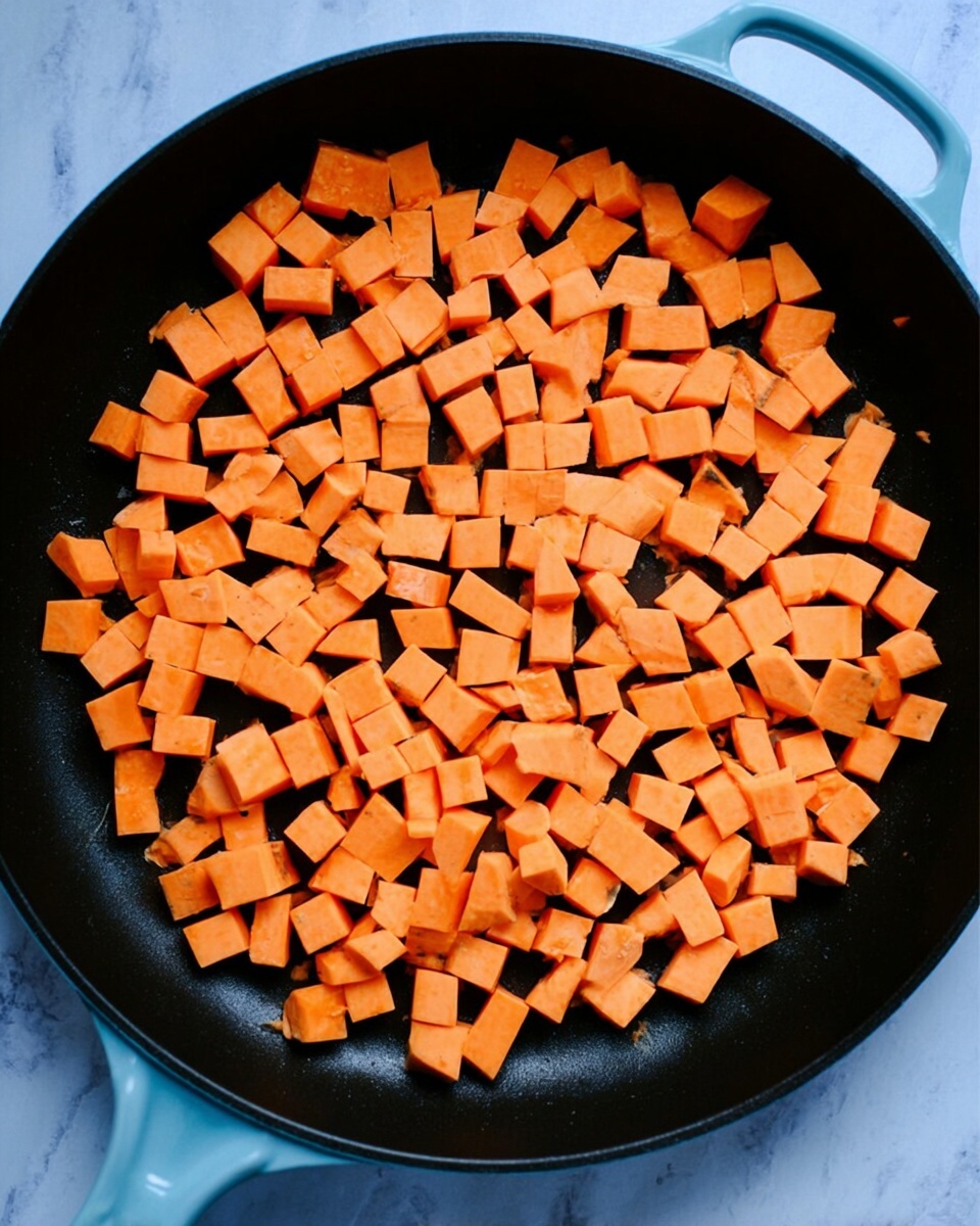 Inside a large black pan with a light blue handle, many small cubes of bright orange sweet potatoes are spread out evenly, covering the entire bottom of the pan. The pan sits on a white marbled surface, creating a clean and simple background. The sweet potatoes' smooth texture and vibrant color stand out clearly against the dark pan and light backdrop. photo taken with an iphone --ar 4:5 --v 7