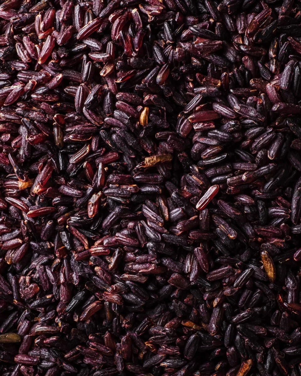 This image shows a close-up of cooked black or purple rice filling the entire frame. The grains are moist, shiny, and tightly packed together, showing a mix of dark purples, deep browns, and black colors with a slightly sticky texture. The surface has a subtle gloss, highlighting the softness and slight fluffiness of the rice. There is no plate or background visible, just the rich, textured rice as the main focus. Photo taken with an iphone --ar 4:5 --v 7