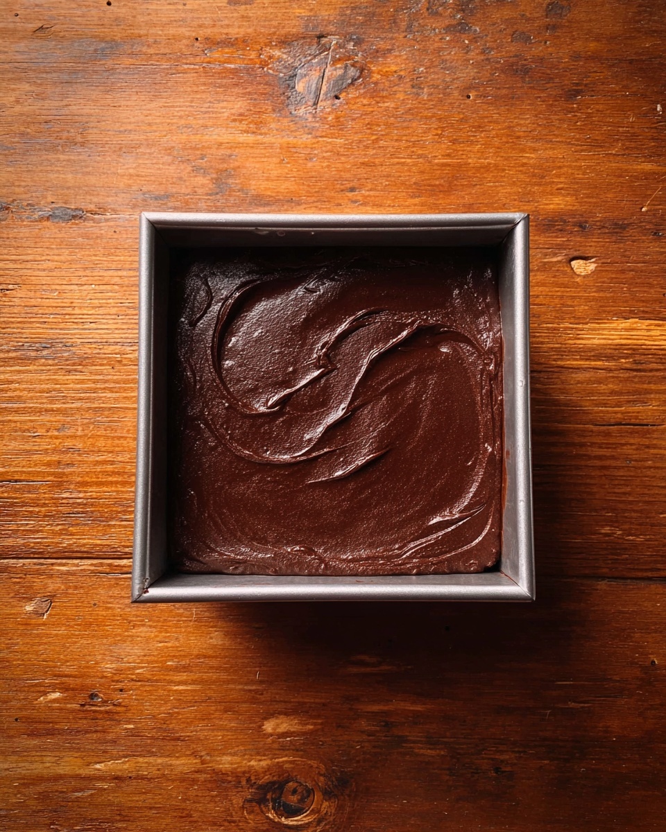 The image shows a square metal baking pan filled with a smooth, thick, dark brown chocolate batter. The surface of the batter has a slightly shiny texture with swirled patterns, indicating it has been spread evenly inside the pan. The pan is placed on a wooden table with a warm, rich brown color and visible grain and knots. photo taken with an iphone --ar 4:5 --v 7