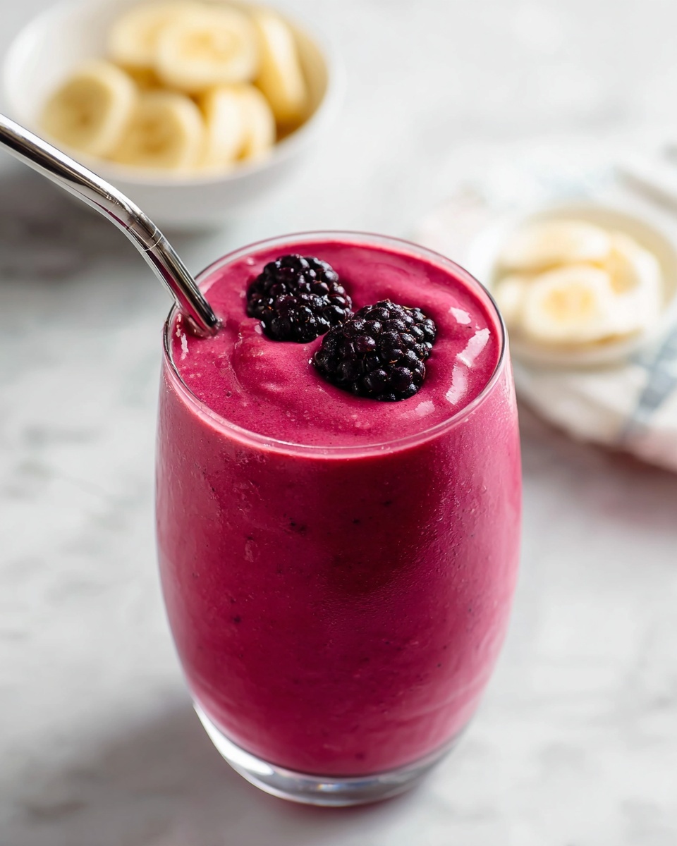 A smooth bright pink smoothie fills a clear round glass, almost to the top, showing a thick texture with small bubbles on the surface and sides. On top of the smoothie, two dark purple-black blackberries sit as a garnish, adding a bumpy texture. A shiny metal straw leans against the glass, slightly bent near the top. The glass rests on a white marbled surface with a soft blurred background that keeps focus on the drink. photo taken with an iphone --ar 4:5 --v 7