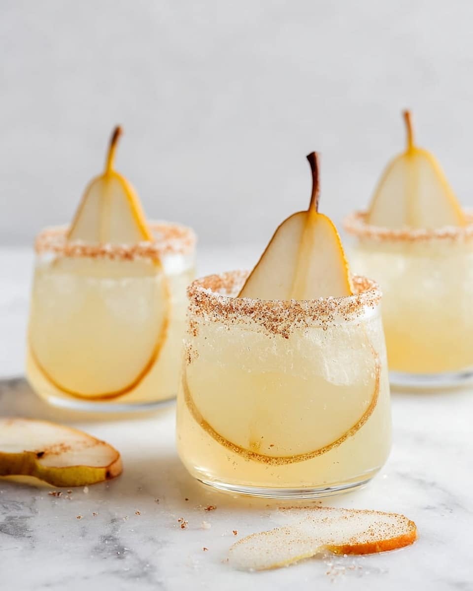 The image shows three clear glasses filled with a light yellow drink. Each glass has a thin round slice of pear inside, with the pear stem sticking out of the glass closest to the front. The rims of the glasses are coated with a mix of sugar and cinnamon, giving a slightly grainy texture around the edge. The glasses sit on a white marbled surface with a few pear slices scattered nearby. The background is plain and light, making the drinks stand out. photo taken with an iphone --ar 4:5 --v 7