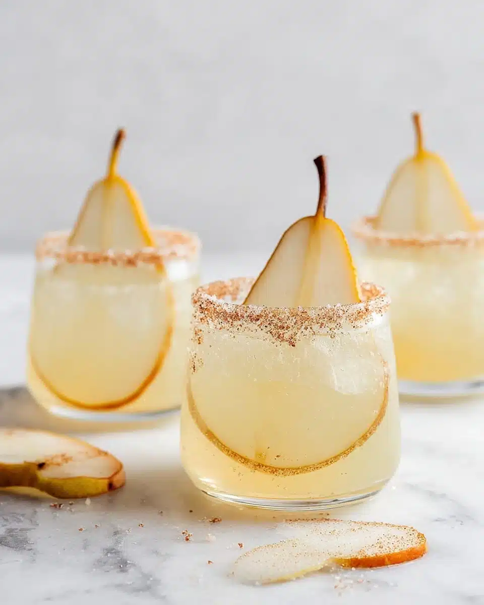 The image shows three clear glasses filled with a light yellow drink. Each glass has a thin round slice of pear inside, with the pear stem sticking out of the glass closest to the front. The rims of the glasses are coated with a mix of sugar and cinnamon, giving a slightly grainy texture around the edge. The glasses sit on a white marbled surface with a few pear slices scattered nearby. The background is plain and light, making the drinks stand out. photo taken with an iphone --ar 4:5 --v 7