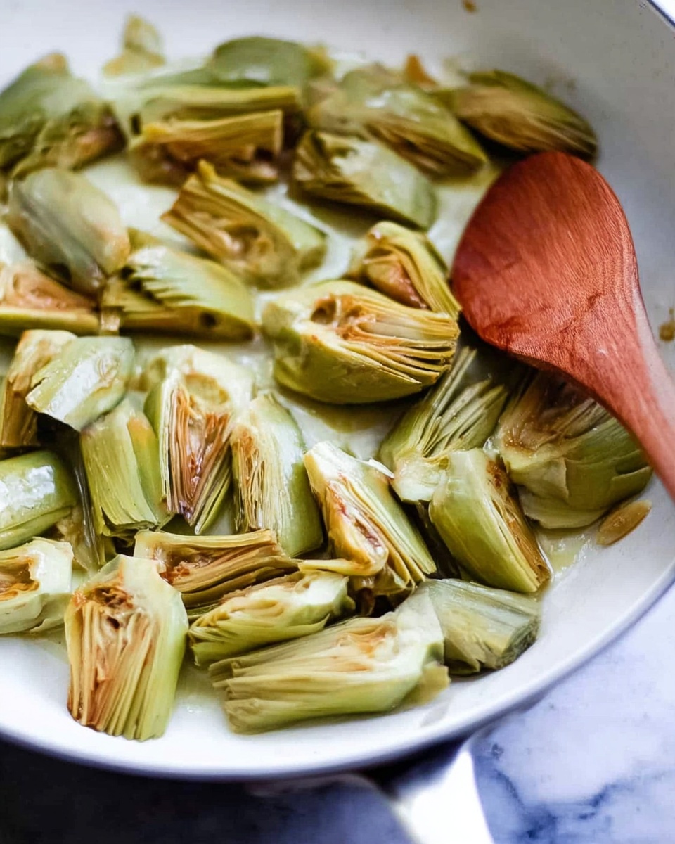 The image shows a white pan with cut artichoke pieces that are light green with some brown edges, indicating they have been cooked. The artichoke pieces are spread out across the pan's surface, some overlapping each other. A wooden spoon with a smooth, brown surface rests on top of the artichokes inside the pan. The background is a white marbled texture. photo taken with an iphone --ar 4:5 --v 7