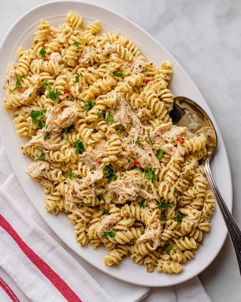 The dish shows a large white oval plate filled with creamy rotini pasta mixed with shredded light chicken pieces. The pasta is coated evenly with a pale yellow sauce speckled with black pepper and small red bits. Fresh green parsley leaves are scattered on top, adding a bright contrast. A large silver serving spoon rests on the right side of the plate. The plate is placed on a white marbled surface with a white cloth having thin red stripes at the bottom left corner. Photo taken with an iphone --ar 4:5 --v 7