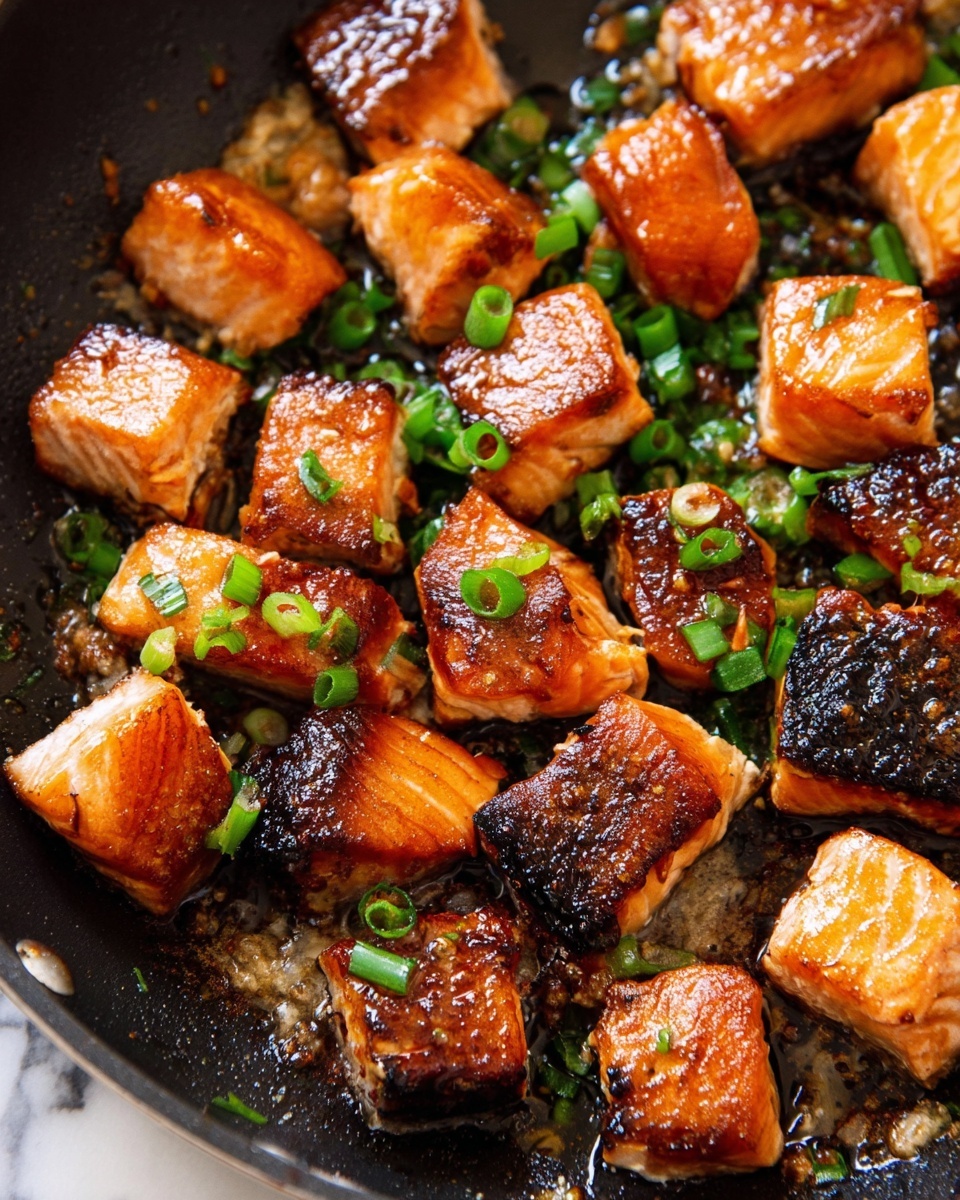 This image shows many small, shiny golden-brown salmon chunks cooked with crispy edges and tender inside. Some pieces have dark crispy skin on one side. Bright green chopped scallions are scattered on top, adding fresh color contrast. The salmon sits in a dark frying pan with a little bit of light oil and cooked bits around the fish pieces. The background is white marbled texture. photo taken with an iphone --ar 4:5 --v 7