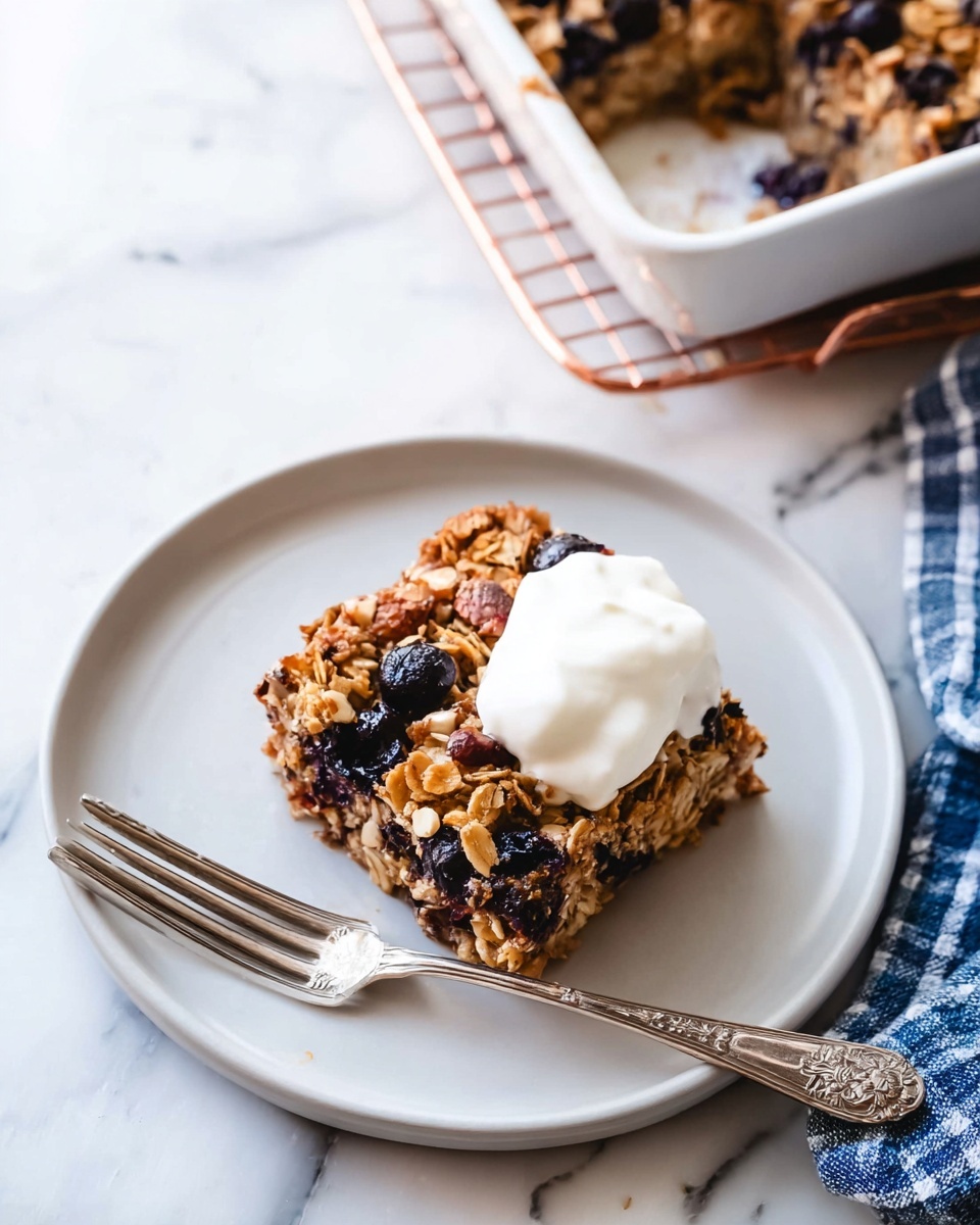 A square piece of baked oatmeal sits near the middle of a round white plate, with layers showing a golden brown oat base mixed with dark blueberries and small nut pieces. On the right side of the oatmeal square, there is a dollop of white creamy yogurt that looks soft and smooth. A silver fork with an ornate handle rests on the plate below the yogurt, pointing diagonally to the bottom left. In the background, the top right corner shows a white baking dish with more blueberry oat bake inside on a round copper cooling rack. The whole scene is on a white marbled surface with a folded blue and white checkered cloth visible on the right edge. photo taken with an iphone --ar 4:5 --v 7