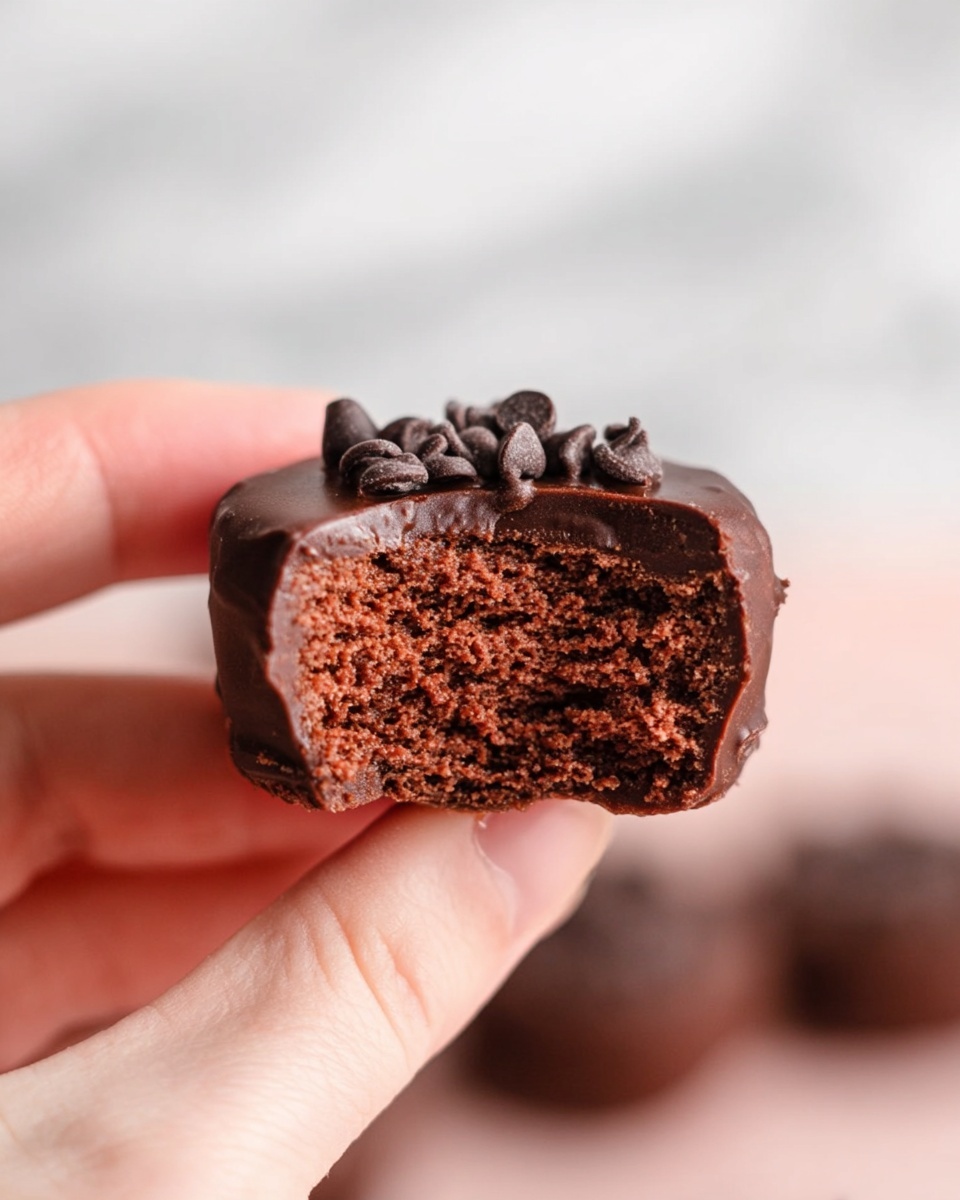 A white bowl filled with multiple round chocolate balls, each coated in smooth dark chocolate and topped with small chocolate chips. The balls are stacked closely together, showing a shiny, slightly textured chocolate surface with scattered chocolate chips on top. The bowl sits on a white marbled surface, with small chocolate chips scattered around it. A pink and white checkered cloth and a glass bottle with white liquid are partially visible in the background. Photo taken with an iphone --ar 4:5 --v 7