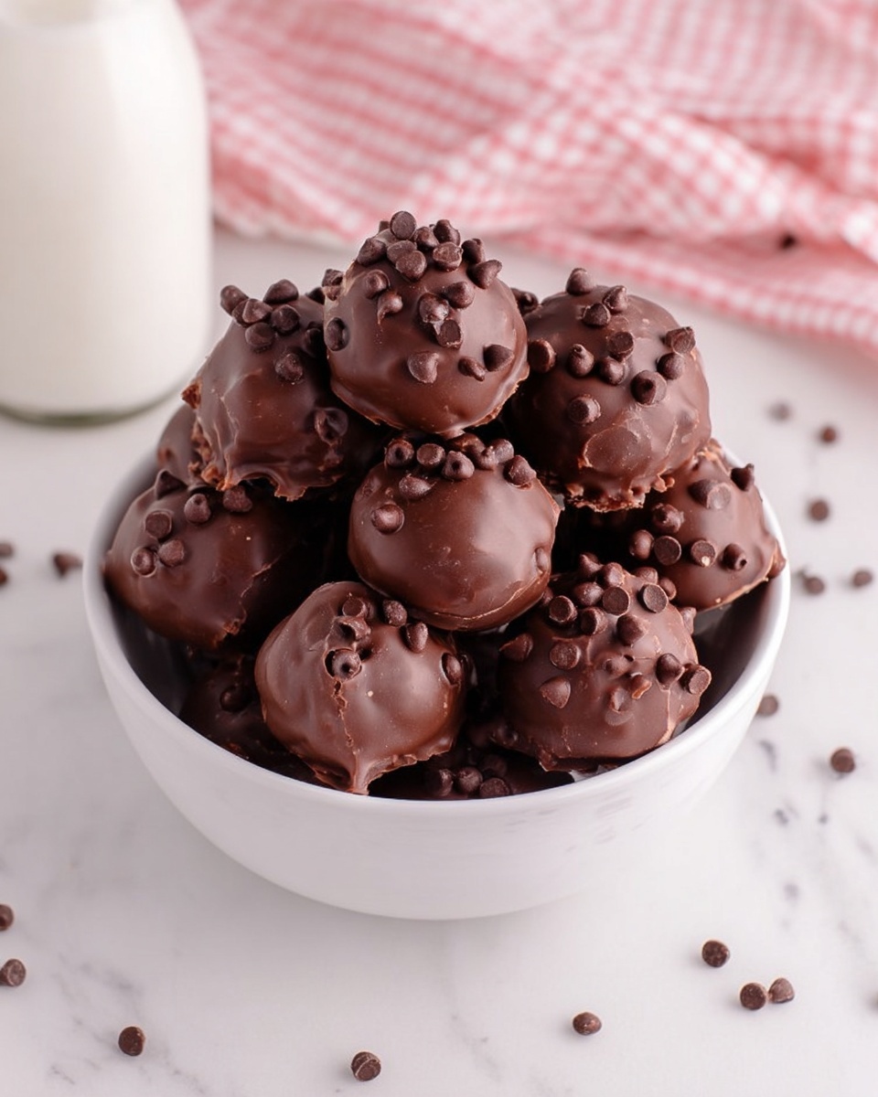 A close-up view of a small round chocolate treat held between a woman's thumb and forefinger, showing three layers: the outer dark chocolate shell, a rich brown dense cake-like middle layer with a moist texture, and small dark chocolate chips scattered on top of the shell. The background features a white marbled texture softly blurred, with a hint of more chocolate treats visible below. The overall colors are dark brown and chocolate tones with natural skin tones of the woman's hand. photo taken with an iphone --ar 4:5 --v 7