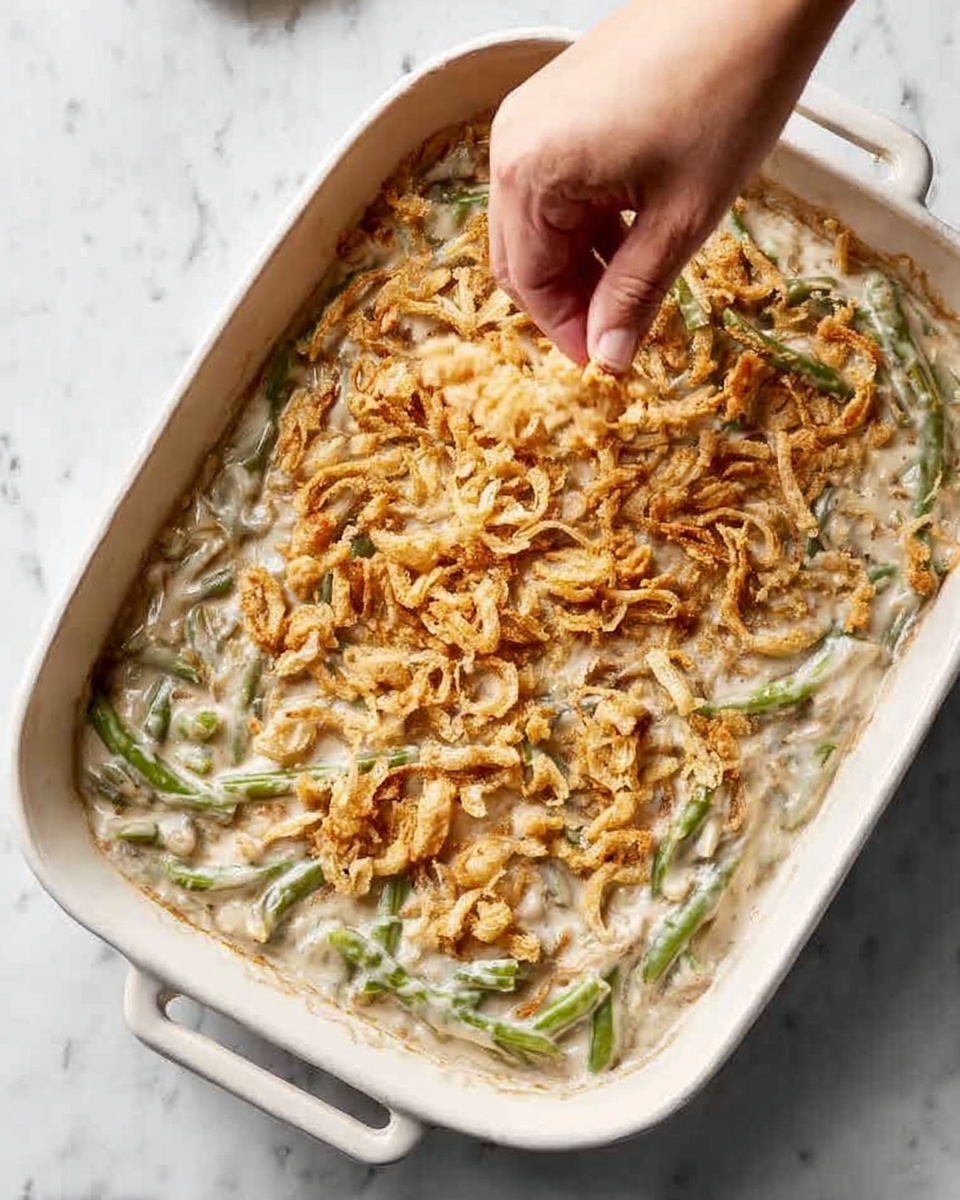 A white rectangular baking dish filled with a creamy, textured green bean casserole that has a light beige sauce covering the green beans underneath. On top, there is a layer of golden, crispy fried onions being sprinkled by a woman's hand coming from the upper left corner. The dish sits on a white marbled surface. Photo taken with an iphone --ar 4:5 --v 7