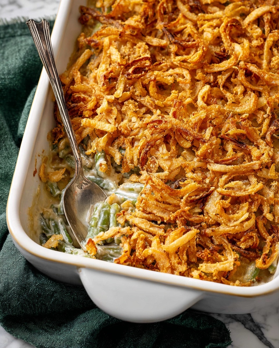 This image shows a white rectangular baking dish filled with a creamy green bean casserole topped with a thick layer of crispy golden-brown fried onions. The casserole itself appears to have a pale, creamy sauce visible underneath the crunchy onion topping, with some green bean shapes peeking through in a few spots. A silver serving spoon is placed inside the dish, slightly scooping a portion from one corner, revealing a smooth, soft layer below the onions. The dish rests on a white marbled surface with a dark green cloth partially visible under one side. Photo taken with an iphone --ar 4:5 --v 7