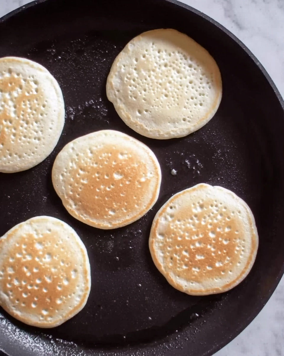 The image shows a close-up of a black round pan with five small pancakes cooking on it. Each pancake is light golden color with many small bubbles on the surface, indicating they are in the process of cooking. The pancakes are evenly spaced across the pan. The background is a white marbled texture. Photo taken with an iphone --ar 4:5 --v 7