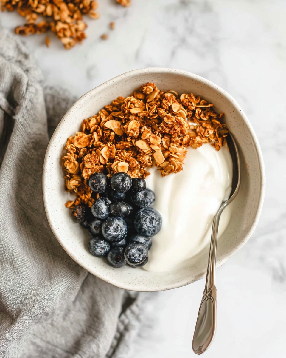 A white bowl filled with three layers: the bottom layer is smooth white yogurt, topped on one side with dark blue blueberries, and on the other side with a heap of golden brown granola that has visible oats and small nuts; a silver spoon rests inside the bowl on the right side, all placed on a white marbled surface with a soft grey cloth nearby. photo taken with an iphone --ar 4:5 --v 7