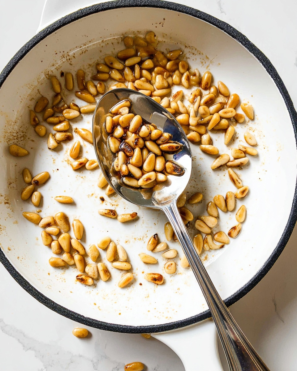 The image shows a white pan with a black rim, containing many small, shiny toasted pine nuts scattered around. A silver spoon, resting inside the pan, holds a small pile of the golden-brown pine nuts, some darker toasted than others. The pan's white surface has light brown spots, showing where the nuts were toasted. The background is a white marbled texture. photo taken with an iphone --ar 4:5 --v 7