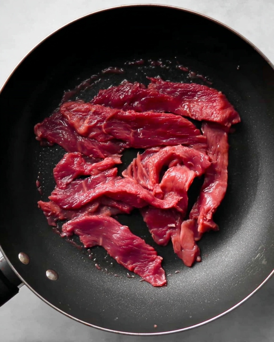 Thin red raw meat strips are spread unevenly in the middle of a black frying pan, with some pieces layered on top of each other and others lying flat around the edges. The meat texture appears soft and slightly moist, showing some natural folds and slight marbling. The black pan has a smooth surface with small droplets of liquid near the bottom. The whole pan is placed on a white marbled surface. photo taken with an iphone --ar 4:5 --v 7