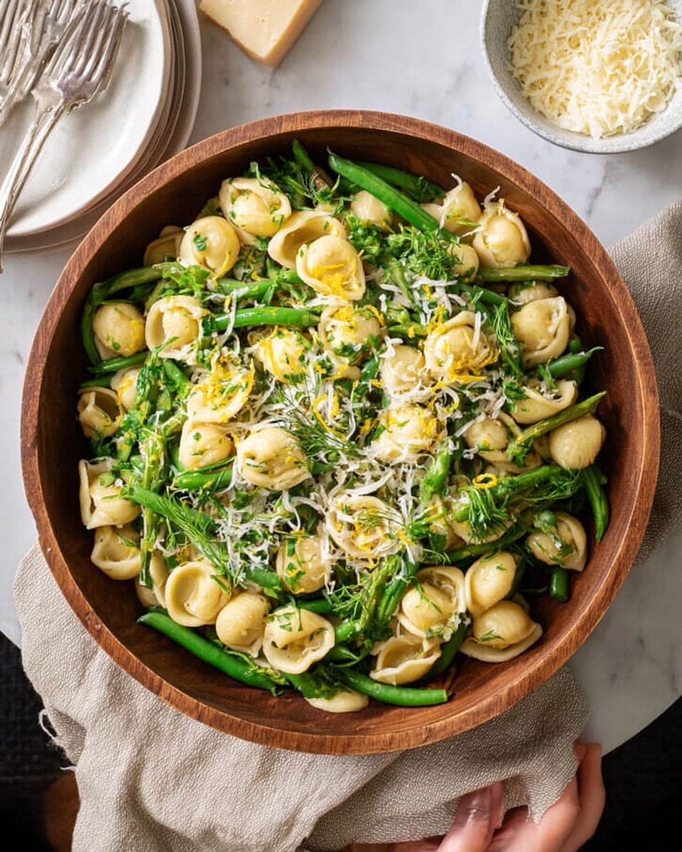 A round dark wooden bowl filled with a layered pasta dish. The bottom layer is green beans, bright and fresh, arranged vertically and spread evenly across the bowl. On top of the green beans are light yellow pasta shells, scattered all around, mixed gently with green leafy herbs. A light dusting of finely shredded white cheese covers the whole dish, adding a soft texture. Small bits of lemon zest are sprinkled on top, giving tiny touches of bright yellow. The bowl is placed on a white marbled surface with a folded beige cloth underneath. Nearby are stacked white plates with silver forks, and a white bowl filled with grated cheese. A woman's hand is reaching out near the plates. photo taken with an iphone --ar 4:5 --v 7