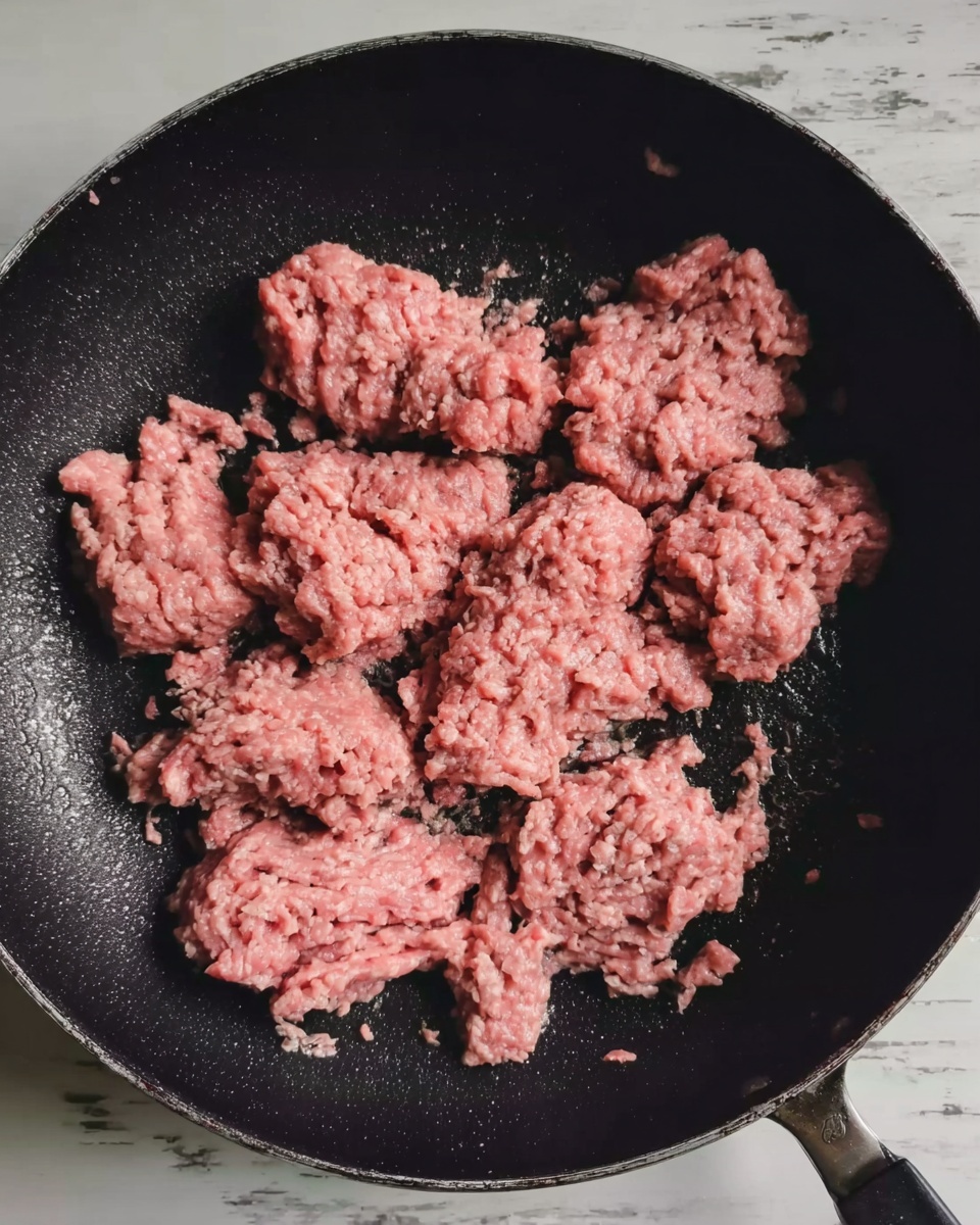 The image shows a black frying pan with several uneven clumps of raw ground meat spread around inside it. The meat looks pink with a moist texture and some small bits of white fat visible. The pan rests on a white marbled surface, and no other objects or utensils are visible in the frame. Photo taken with an iphone --ar 4:5 --v 7