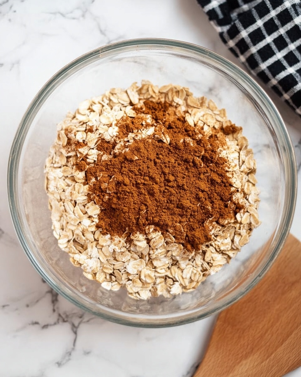 A clear glass bowl is filled with a base layer of light beige rolled oats. On top of the oats is a medium layer of fine brown cinnamon powder, slightly spread but mostly centered. The bowl sits on a white marbled surface, with part of a black and white checkered cloth visible on the upper right side. A wooden spatula lies just below the bowl, partially visible. Photo taken with an iphone --ar 4:5 --v 7