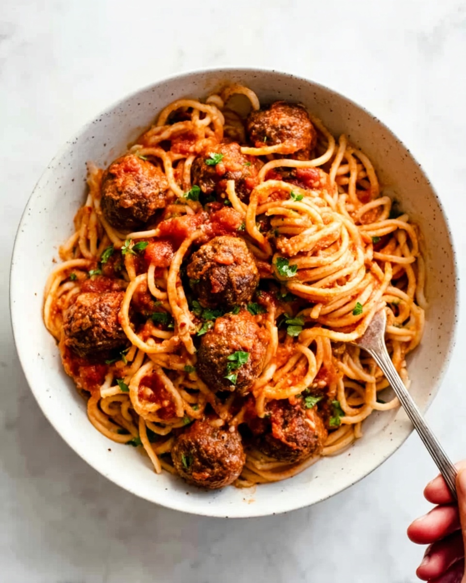 The image shows a close-up of a white plate filled with spaghetti pasta coated in a red tomato sauce mixed with small pieces of tomato and herbs. Scattered evenly among the pasta are several brown meatballs with a slightly rough texture. Fresh green chopped herbs are sprinkled on top, adding a pop of color against the warm tones of the dish. The plate rests on a white marbled surface. photo taken with an iphone --ar 4:5 --v 7