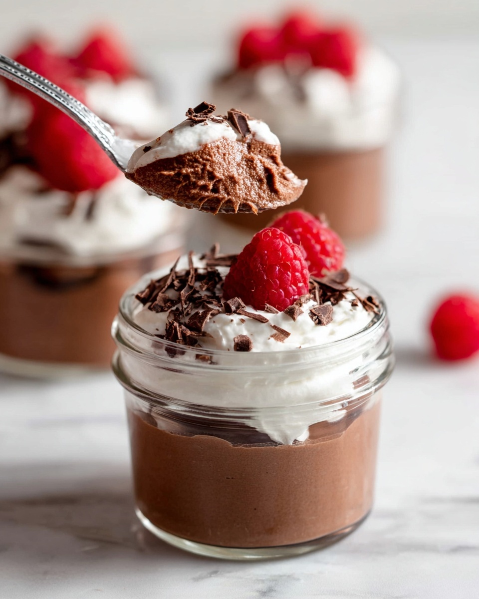 A small clear glass jar is filled with three visible layers: the bottom layer is smooth, dark brown chocolate mousse, the middle layer is topped with fluffy white whipped cream, and the top is decorated with dark chocolate shavings and a bright red raspberry. A silver spoon holds a scoop of the chocolate mousse and whipped cream above the jar. In the blurry background, another jar with similar layers and whipped cream topped with red raspberries sits on a white marbled surface. Photo taken with an iphone --ar 4:5 --v 7