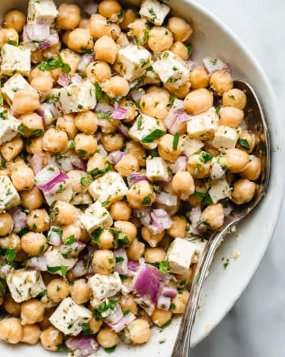 A white bowl filled with a chickpea salad showing three main layers: light orange chickpeas, small white feta cheese cubes, and diced purple-red onion pieces, all mixed with finely chopped green herbs that cover the entire salad. There is a bright yellow lemon wedge placed on the side inside the bowl, and a silver fork is partially dipped in the salad on the right side. The bowl is placed on a white marbled surface, with parts of a lemon and green leaves visible in the top corners, and a striped gray and white cloth is partly visible at the bottom left corner. Photo taken with an iphone --ar 4:5 --v 7