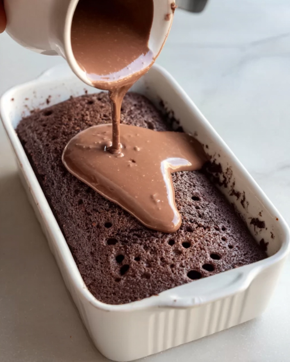 A white rectangular baking dish holds a dark brown chocolate cake with small holes on its surface. Light brown chocolate sauce is being poured over the cake from a white cup, covering the center and dripping down the sides. The background is a white marbled surface, and a woman's hand is visible holding the cup. Photo taken with an iphone --ar 4:5 --v 7