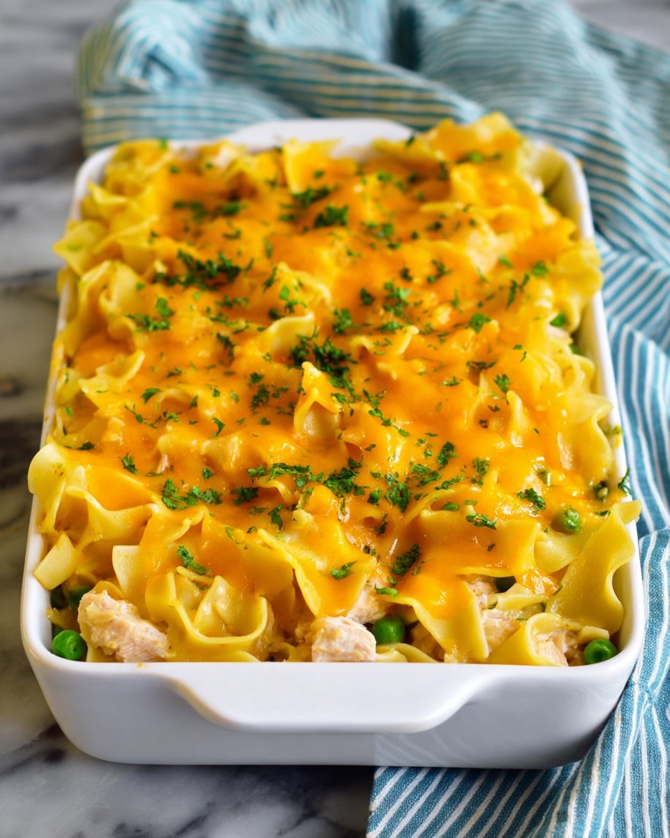 A white rectangular dish filled with a layered pasta casserole. The bottom layer shows wide, flat noodles mixed with small pieces of light-colored cooked chicken and green peas. On top, there is a bright golden-yellow melted cheese layer that covers the pasta evenly, with some green herbs sprinkled across for color contrast. The dish sits on a white marbled surface, and a striped cloth is softly blurred in the background. Photo taken with an iphone --ar 4:5 --v 7
