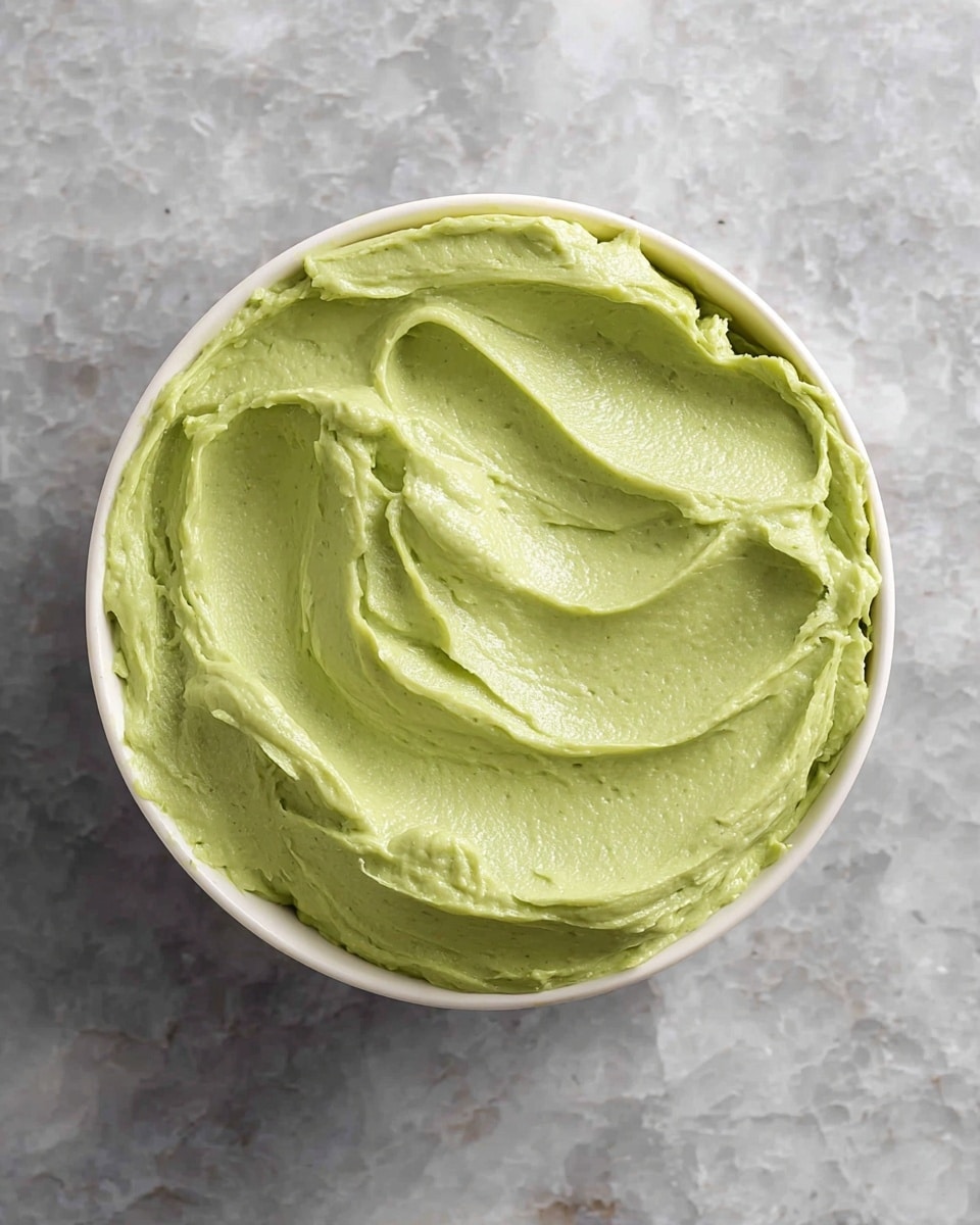A metal rectangular container lined with white parchment paper holds a smooth, pale green ice cream-like mixture with a neat scoop taken from the middle by a silver ice cream scoop resting inside. Around the container on a white marbled surface, there are two halved avocados showing their green flesh and brown seed, along with a few small avocado chunks scattered nearby. The focus is on the creamy texture and fresh avocado colors. photo taken with an iphone --ar 4:5 --v 7