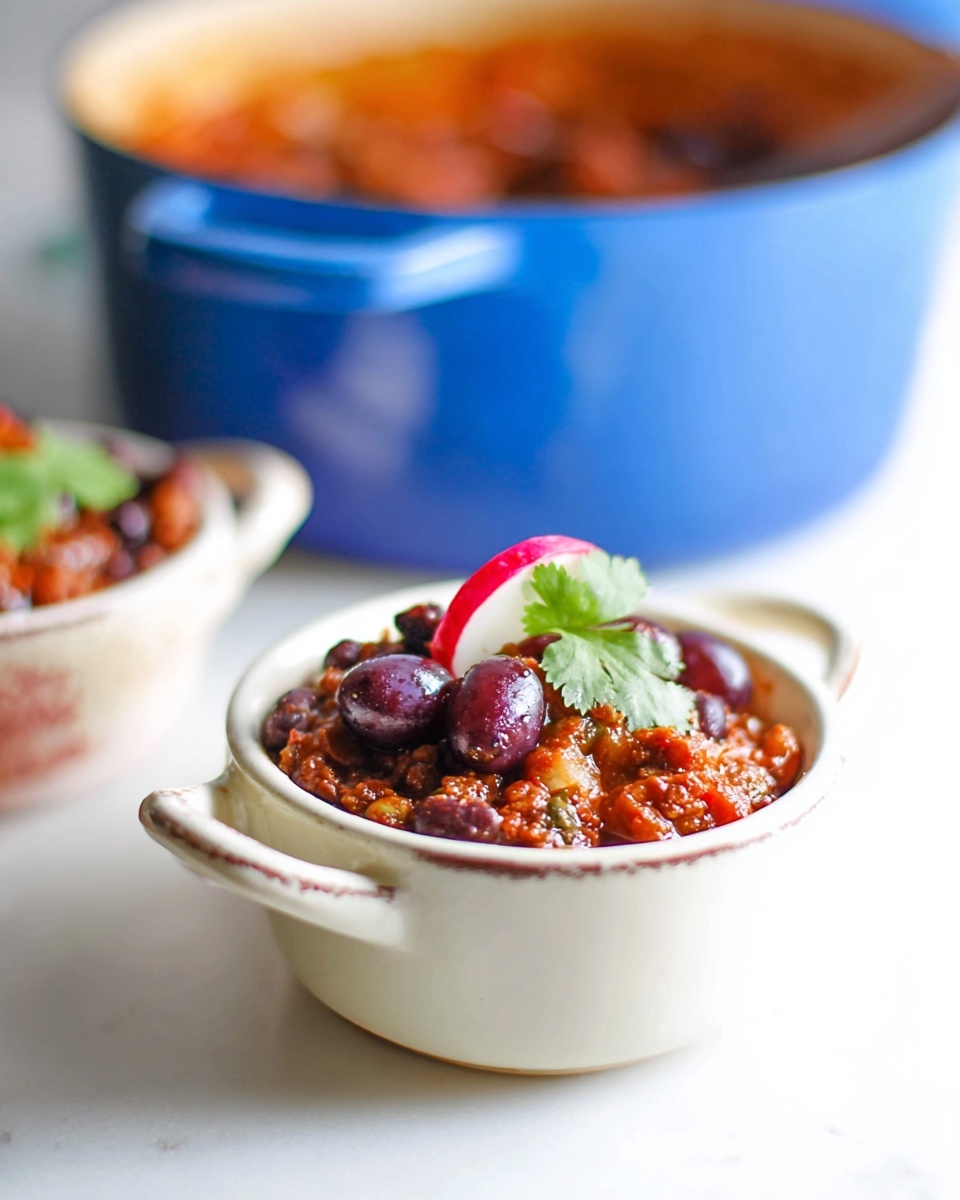 A small white bowl with two handles is filled with a rich, chunky reddish-brown stew that has visible pieces of vegetables and beans, topped with dark purple grapes, a slice of radish, and a small bright green leaf of cilantro. In the background, a large blue pot is slightly out of focus, resting on a smooth white marbled surface. Another small white bowl with handles is also partly visible, containing a similar stew with dark beans on top. The colors are warm and inviting, with a fresh and homey feel. Photo taken with an iphone --ar 4:5 --v 7