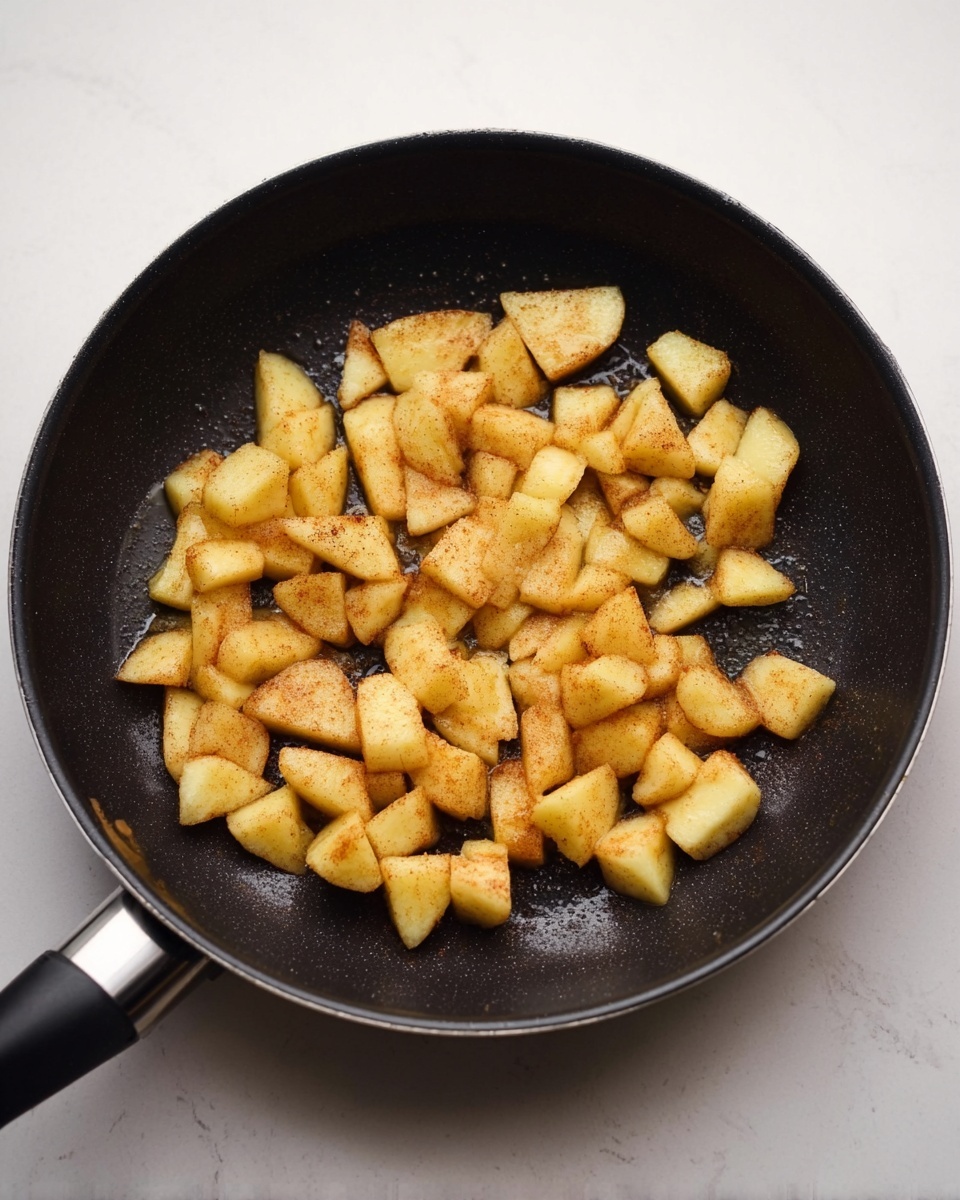 The image shows a black non-stick frying pan filled with small pieces of lightly browned apple slices. The apple pieces are soft yellow with a light dusting of cinnamon, giving them a warm brownish tint. They are spread evenly across the center of the pan, and a little shine suggests they are cooking in some butter or oil. The pan handle extends out to the left side, and the pan sits on a white marbled surface. photo taken with an iphone --ar 4:5 --v 7