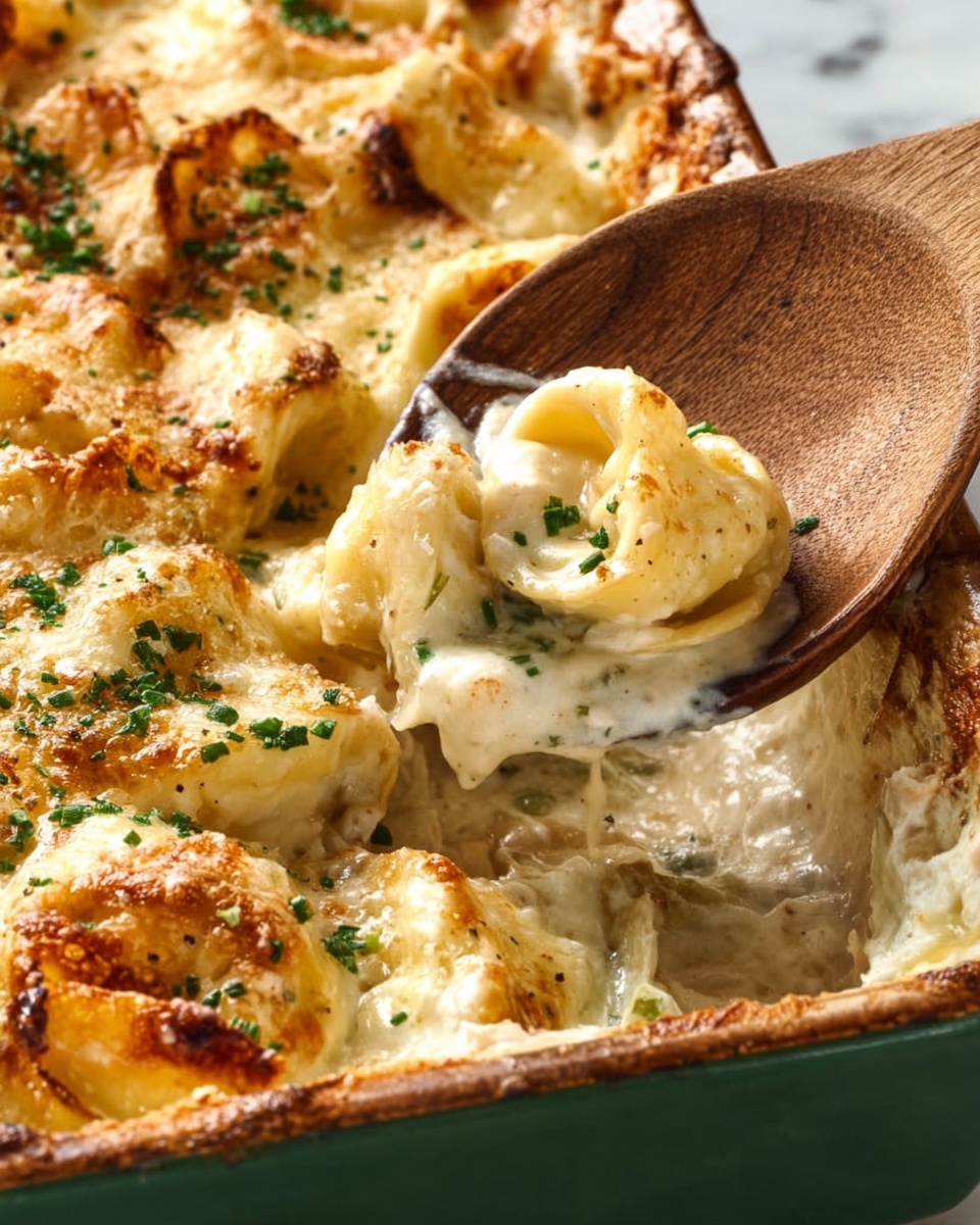 A close-up image of a baked pasta dish in a green ceramic baking dish sitting on a white marbled surface. The dish has a golden-brown cheese layer on top with small green herb flakes scattered over it. Beneath the crispy cheese crust, there are creamy layers of tortellini pasta filled with cheese, coated in a smooth white sauce with a few browned spots. A wooden spoon lifts one tortellini showing its soft, creamy texture with herbs inside. The edges of the baking dish show some baked cheese stuck to the sides. photo taken with an iphone --ar 4:5 --v 7