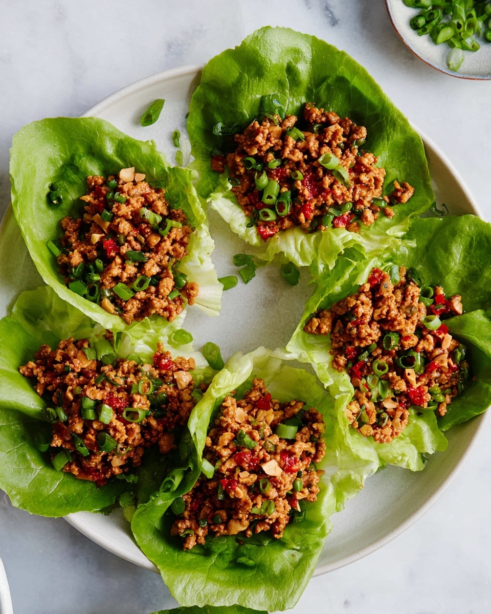 The image shows five light green lettuce cups arranged in a circle on a white plate, each cup filled with a layer of cooked, finely crumbled brown meat mixed with green onion pieces and tiny red chili bits. The meat mixture has a slightly shiny texture with some small chunks of nuts visible throughout. The lettuce leaves are fresh, smooth, and gently curved to hold the filling, contrasting with the textured meat on top. Some chopped green onions are scattered on and around the plate, which sits on a white marbled surface. Photo taken with an iphone --ar 4:5 --v 7