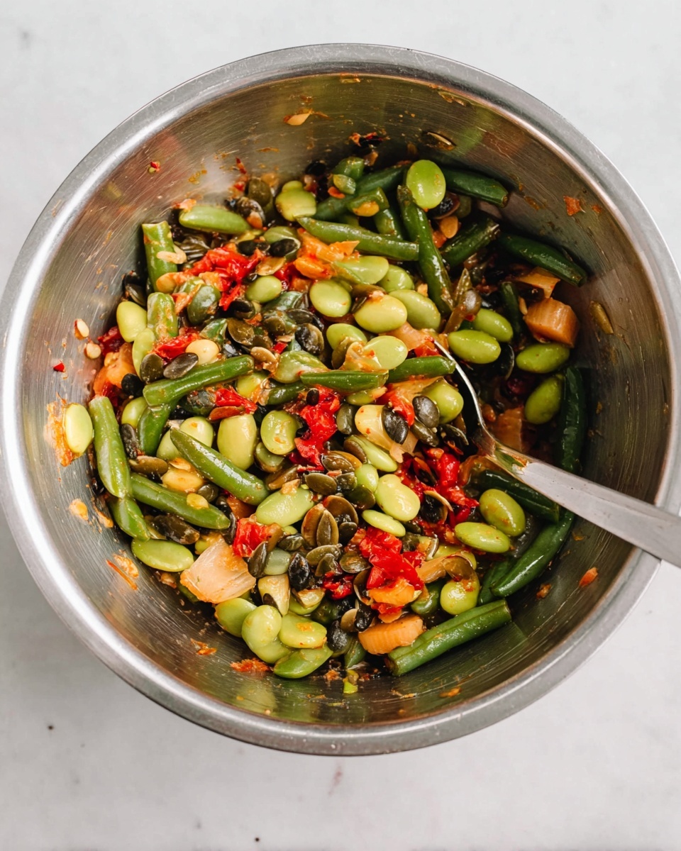 A shiny metal bowl filled with a colorful mix of large, smooth green beans, small dark green pumpkin seeds, and chunk pieces of red and light yellow vegetables, all lightly coated in a glossy dressing. A metal spoon rests inside the bowl on the right side, partially covered by the salad mix. The bowl sits on a white marbled surface. photo taken with an iphone --ar 4:5 --v 7