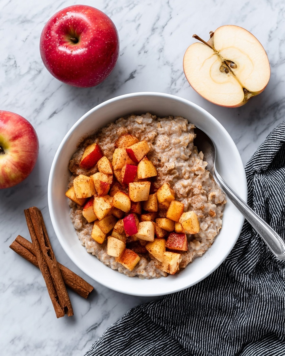 A white bowl filled with a creamy light brown oatmeal base, topped with a generous layer of bright red and yellow apple chunks that have a slightly soft and shiny texture, sprinkled with ground cinnamon giving a speckled look. A silver spoon rests inside the bowl on the left side. The bowl sits on a white marbled surface, with a red apple and two cinnamon sticks placed nearby. Photo taken with an iphone --ar 4:5 --v 7