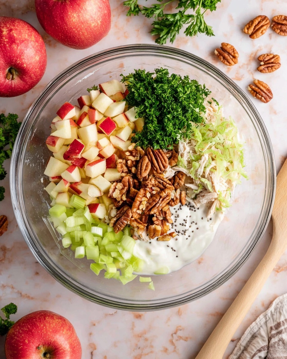 A clear glass bowl holds six separate layers of ingredients arranged neatly in sections inside it. Starting from the top left and moving clockwise, there are small red and white apple cubes, finely chopped bright green parsley, small pieces of shredded light brown cooked chicken, chopped brown pecans with black pepper and some seeds sprinkled over, curved pale green celery slices, and a smooth white creamy layer at the bottom left. The bowl sits on a white marbled surface with red apples and parsley scattered around. A beige spatula with a wooden handle lies near the bowl, and a woman's hand is partly visible at the edge of the frame. Photo taken with an iphone --ar 4:5 --v 7