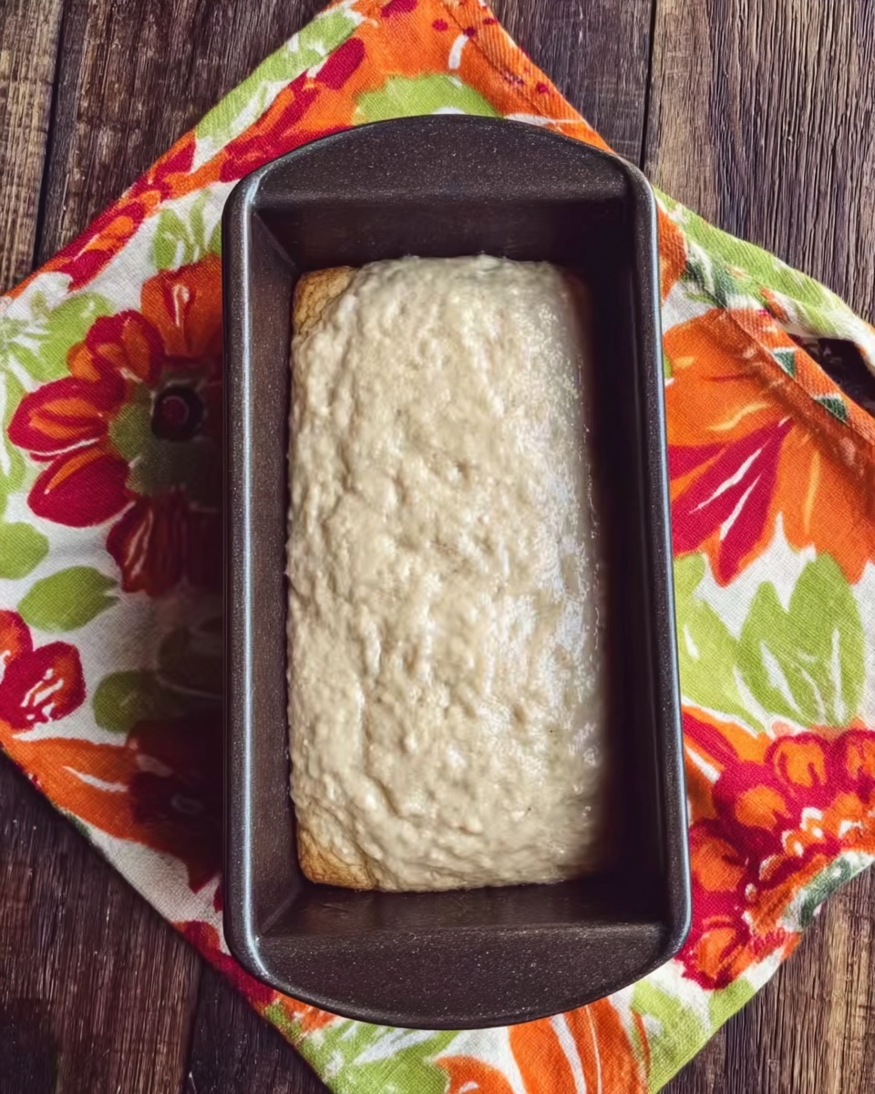 The image shows an unbaked batter inside a rectangular dark metal loaf pan. The batter is light beige in color with a slightly uneven, bumpy texture on the surface. The pan is placed on a colorful cloth with large orange, red, green, and cream floral patterns. The background beneath the cloth is a wooden surface. photo taken with an iphone --ar 4:5 --v 7