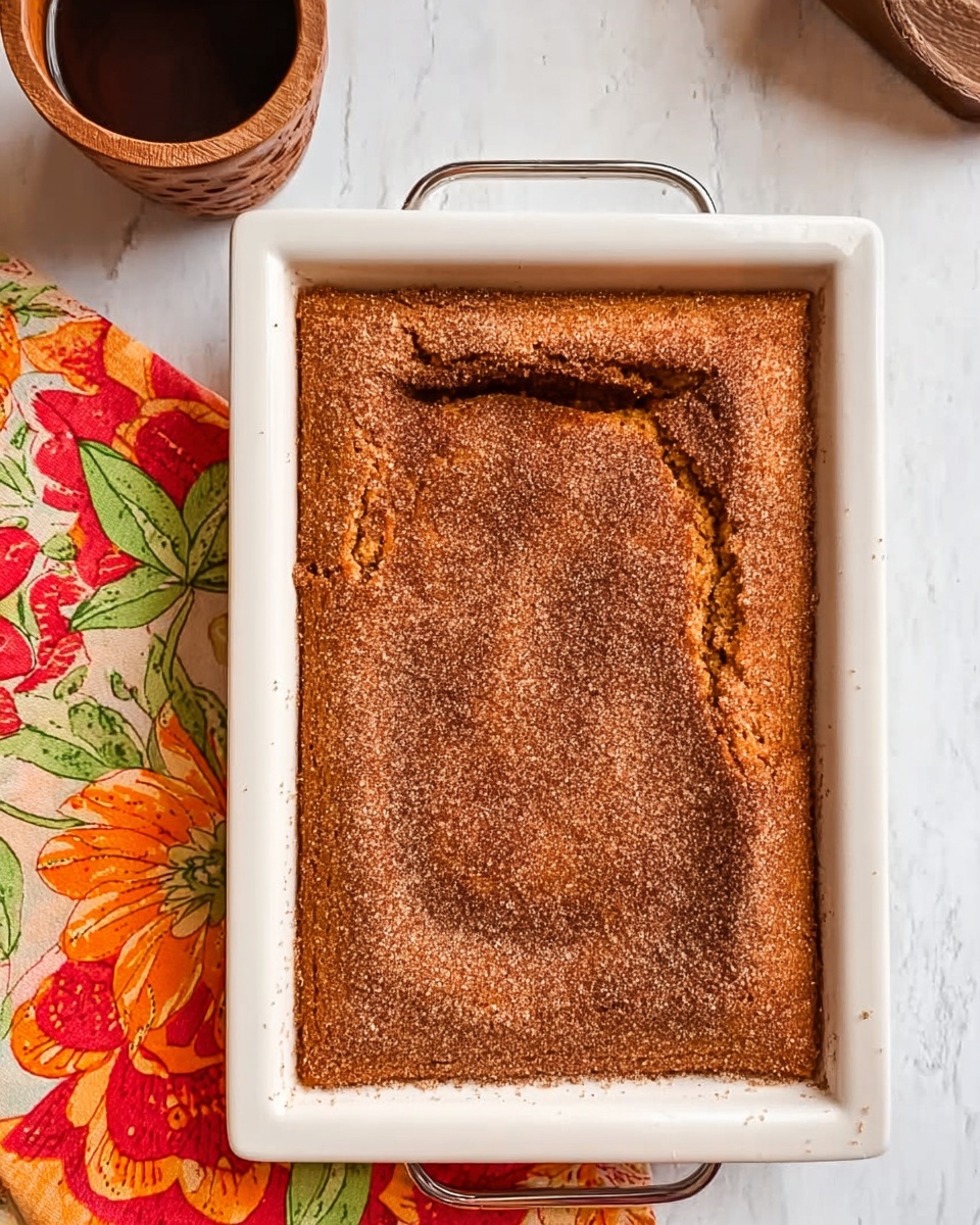A rectangular brown cake with a rough, slightly cracked top layer dusted with sugar sits in a white square pan with a metal handle. The cake surface has a textured look with some darker areas around the edges. It is placed on a white marbled surface with a colorful cloth featuring red, orange, and yellow floral patterns nearby, and a wooden cup is partially visible in the upper left corner. The overall scene is bright and clear. photo taken with an iphone --ar 4:5 --v 7
