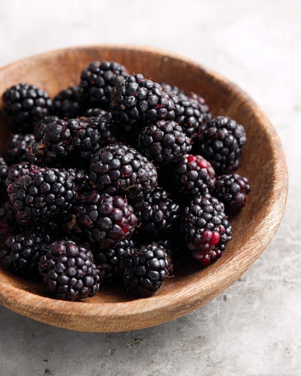 A round wooden bowl filled with many blackberries that are deep purple to black in color with a glossy and bumpy texture, resting on a white marbled surface. photo taken with an iphone --ar 4:5 --v 7