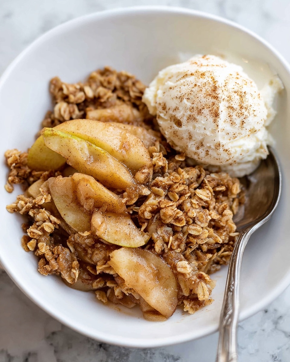 The image shows a white plate with two main parts: on one side, there is a mix of soft, cooked apple slices with a light brown cinnamon color, topped with small clusters of golden oat crumble. On the other side, there is a scoop of creamy white ice cream sprinkled with a light dusting of cinnamon powder. A silver spoon rests on the plate, holding a piece of apple with crumble. The plate is set on a white marbled surface, and in the background, a blurred green apple is visible. photo taken with an iphone --ar 4:5 --v 7