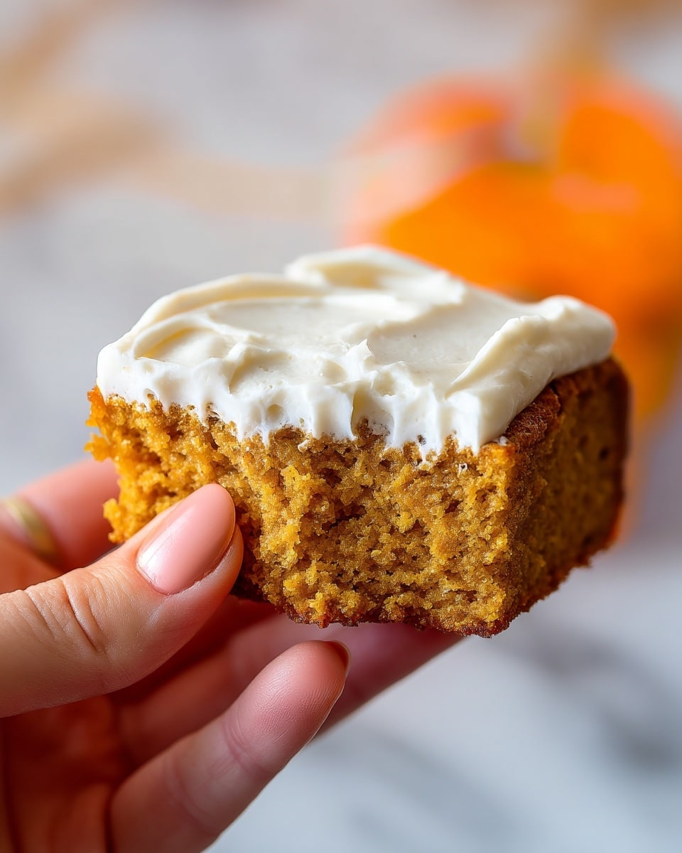 A close-up image of a pumpkin-flavored cake slice held by a woman's hand, showing one bite taken from the bottom center. The cake layer is light brown with a soft and moist texture, topped with a thick, creamy white frosting spread evenly over the surface. The background is a white marbled texture, softly blurred with warm tones hinting at an orange object behind the cake. Photo taken with an iphone --ar 4:5 --v 7