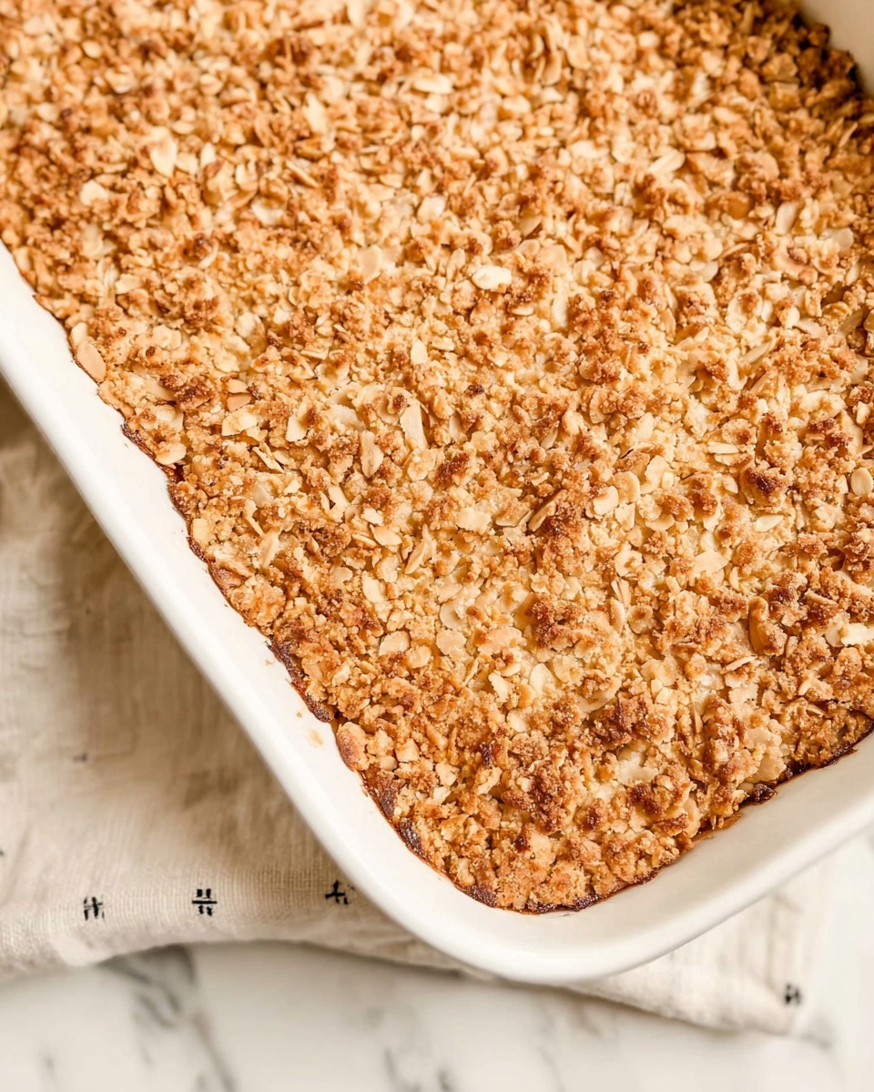 A close-up view of a rectangular white ceramic baking dish filled with a golden-brown topping made of small, crunchy pieces that look like chopped nuts and oats, covering the entire surface evenly. The dish is placed on a white marbled surface with a light beige cloth underneath that has small black cross patterns. The texture of the crunchy topping looks slightly rough and toasted, showing different shades of brown with some darker bits scattered throughout. Photo taken with an iphone --ar 4:5 --v 7