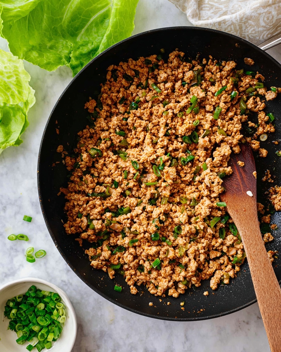 In a black pan filled mainly with light brown cooked ground meat mixed with small, finely chopped green onions and tiny pale yellow bits, the mixture has an even texture and color with slight reddish highlights from the sauce. A wooden spatula rests on the left side inside the pan, partially covered by the food. In the top left corner, bright green lettuce leaves are visible on a white marbled surface, scattered with a few small pieces of green onion. In the bottom right corner, a small white bowl holds bright green chopped herbs or scallions, sitting on the same white marbled surface. Photo taken with an iphone --ar 4:5 --v 7