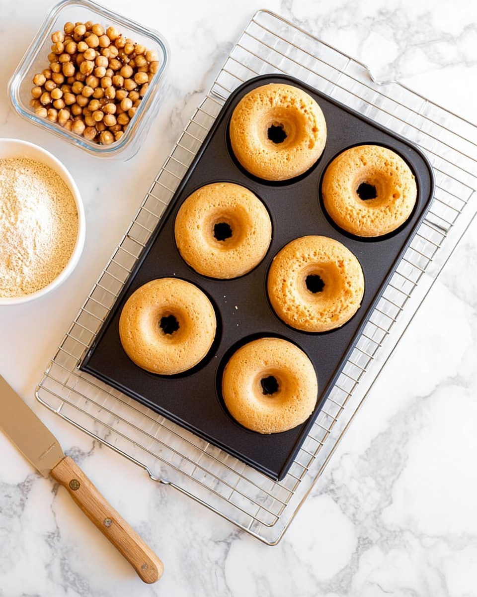 A black metal baking tray holds six freshly baked plain donuts with a light golden brown color and smooth texture. The tray rests on a silver cooling rack placed on a white marbled surface. To the left, there is a clear container with round brown chickpeas and a white bowl filled with light brown flour. A wooden butter knife lies on the surface beside the baking tray. The scene is bright and clean with soft natural lighting. photo taken with an iphone --ar 4:5 --v 7