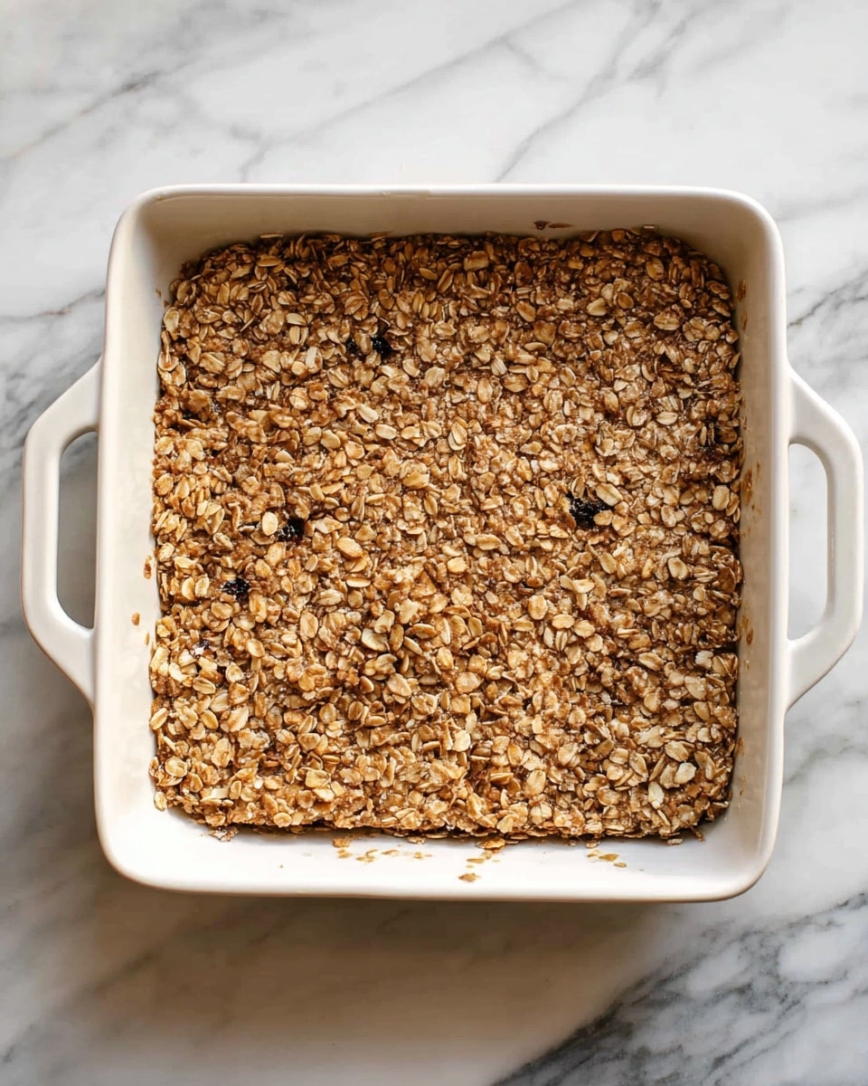 A square white baking dish with small handles on two sides holds a single thick layer of baked oat mixture with a rough texture. The top layer is golden-brown with visible whole oats and small dark bits scattered unevenly throughout, creating a crunchy, crumbly appearance. The dish is placed on a white marbled surface with grey veins. Photo taken with an iphone --ar 4:5 --v 7