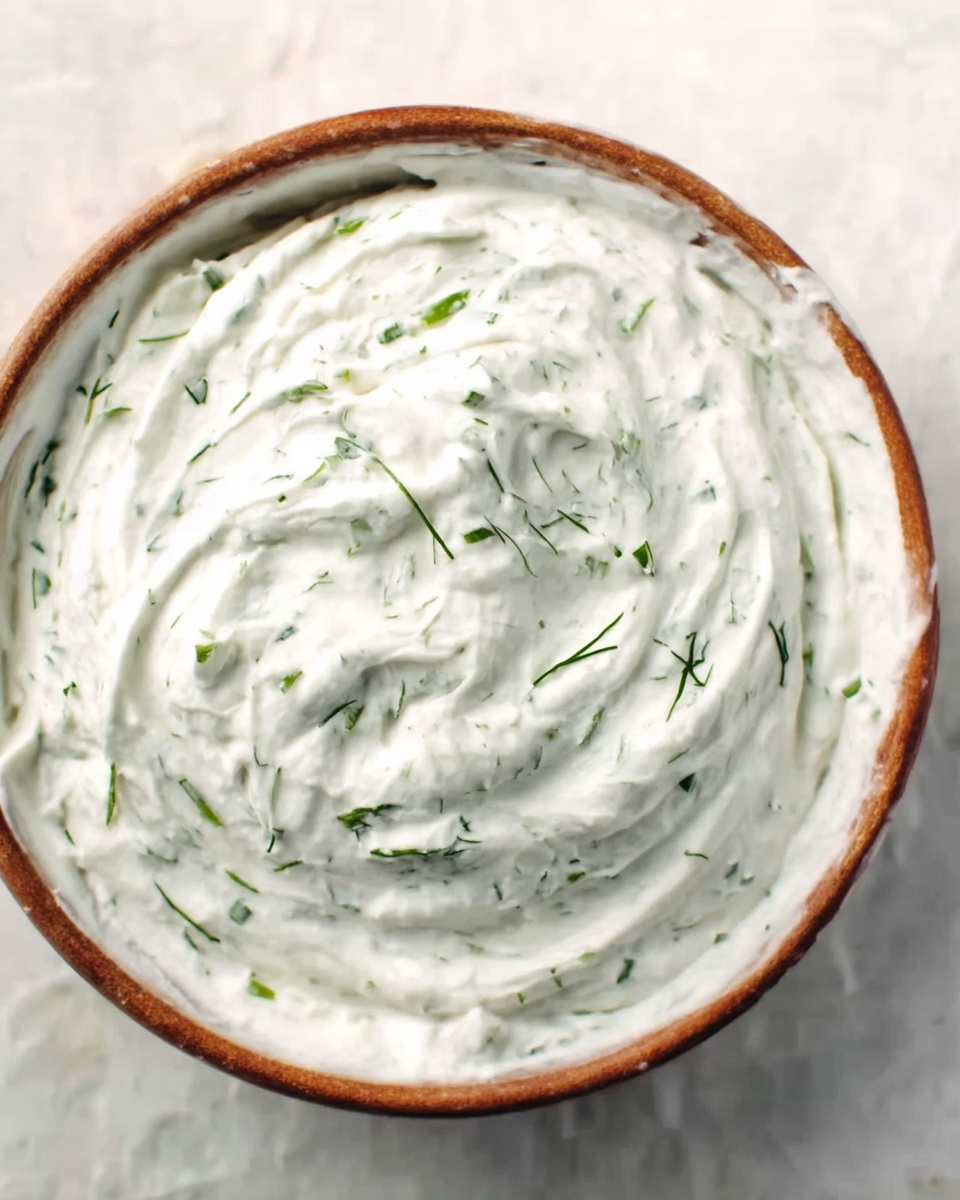 A white marbled textured background holds a round white bowl filled with a thick, creamy white dip. The dip has small bright green herb pieces evenly mixed throughout, giving a fresh look. The surface of the dip shows soft, smooth swirls and some small peaks, highlighting the creamy texture. Photo taken with an iphone --ar 4:5 --v 7